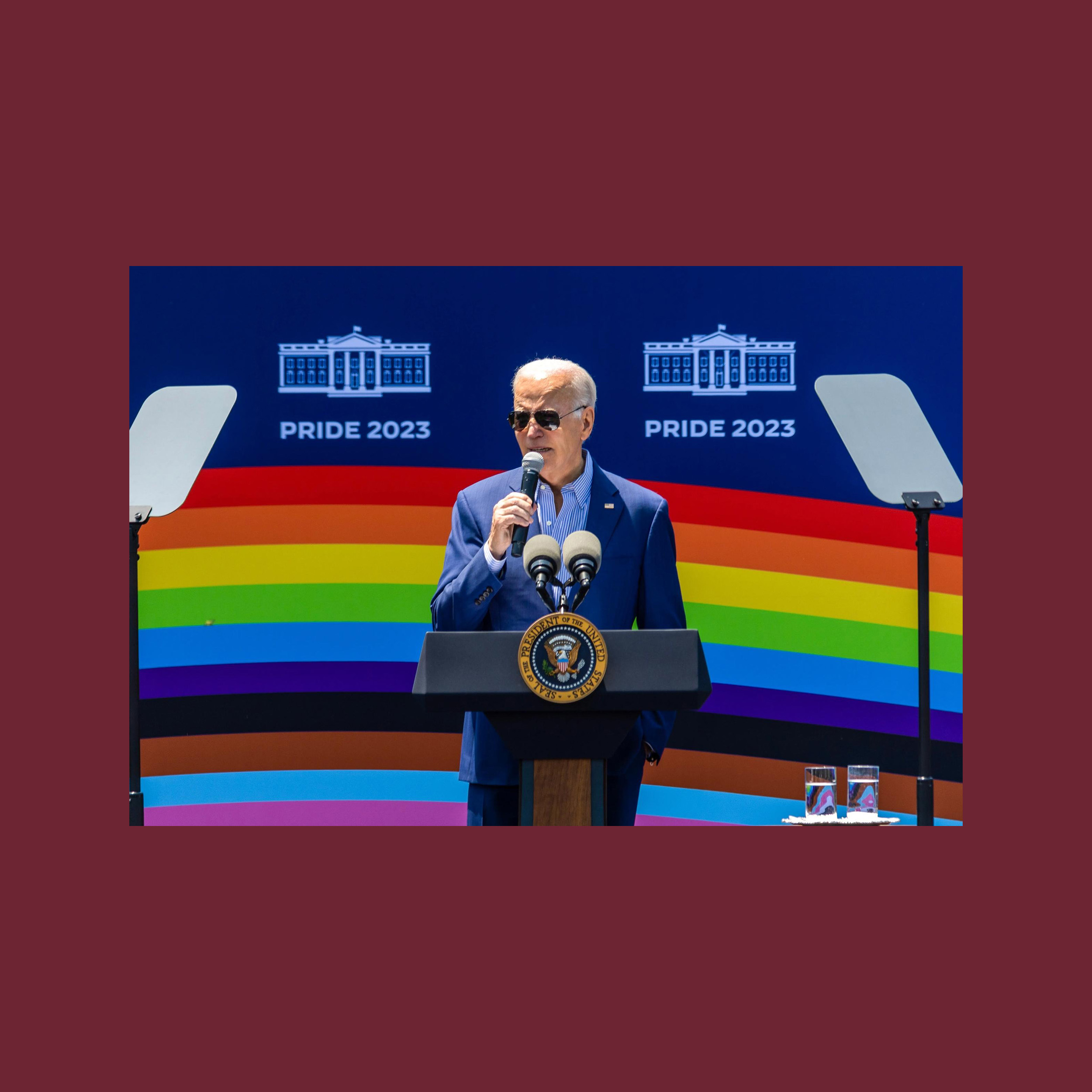 joe biden speaking on a podium in front of a pride rainbow